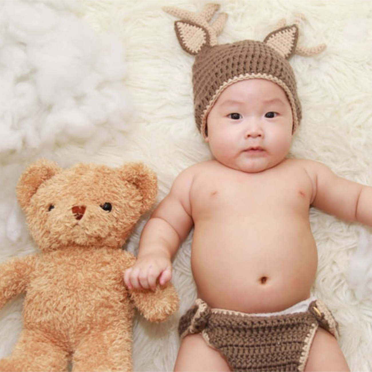 baby boy with knitted diaper and deer hat holding onto stuffed bear.